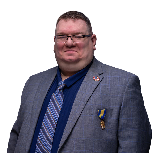 A man wearing glasses, a blue shirt, striped tie, and gray plaid suit jacket with a pin and a medal on the lapel, stands against a plain white background, smiling slightly.